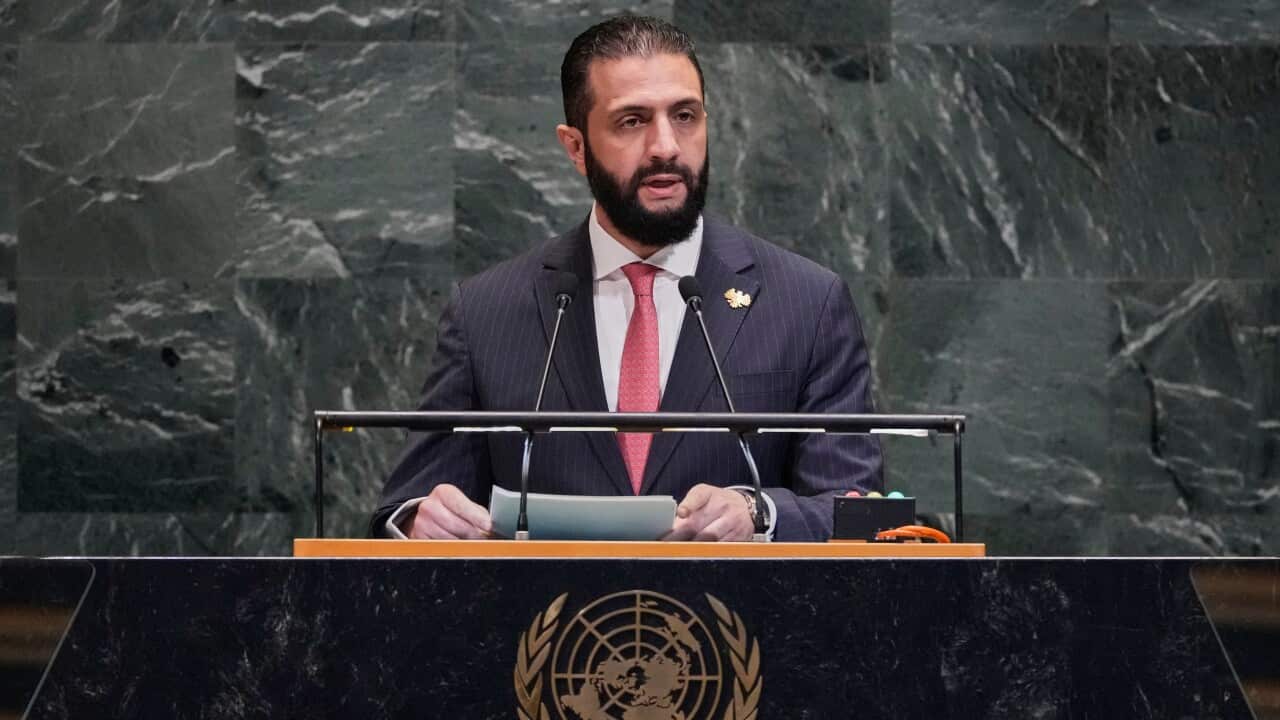 A man in a dark suit and red tie talks from behind a lectern. There is a marble wall behind him