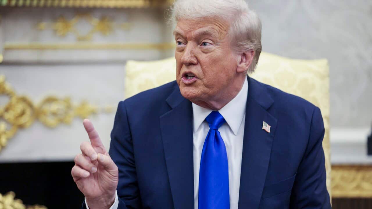Donald Trump, wearing a dark blue suit and a bright blue tie, is pictured from the chest up, gesturing with an upraised index finger as he speaks. A gold-trimmed fireplace and a yellow armchair are visible behind him.