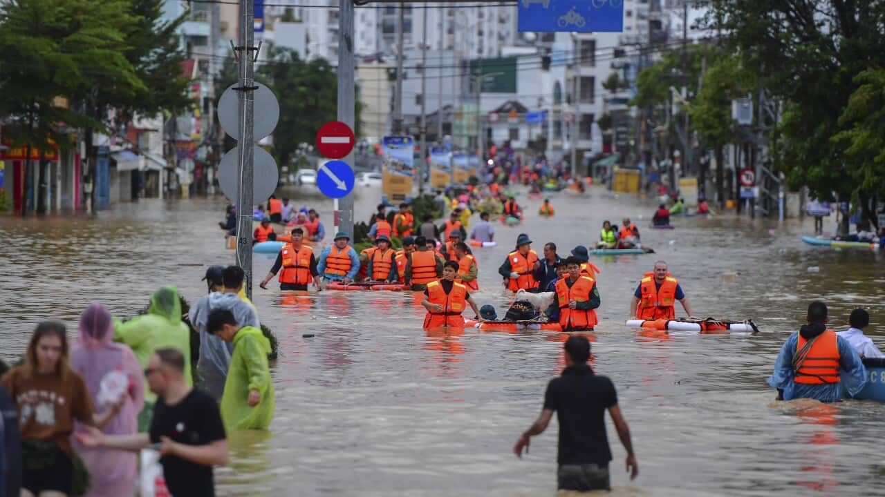 Dozens of people, some wearing high vis life jackets, wading through waist heigh brown flood waters on a city street