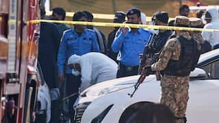 Police and miltary investigate the scene of a suicide bombing in Pakistan. A person in white protective clothing and a mask looks over a burnt out car. Police and soliders holding guns stand nearby.