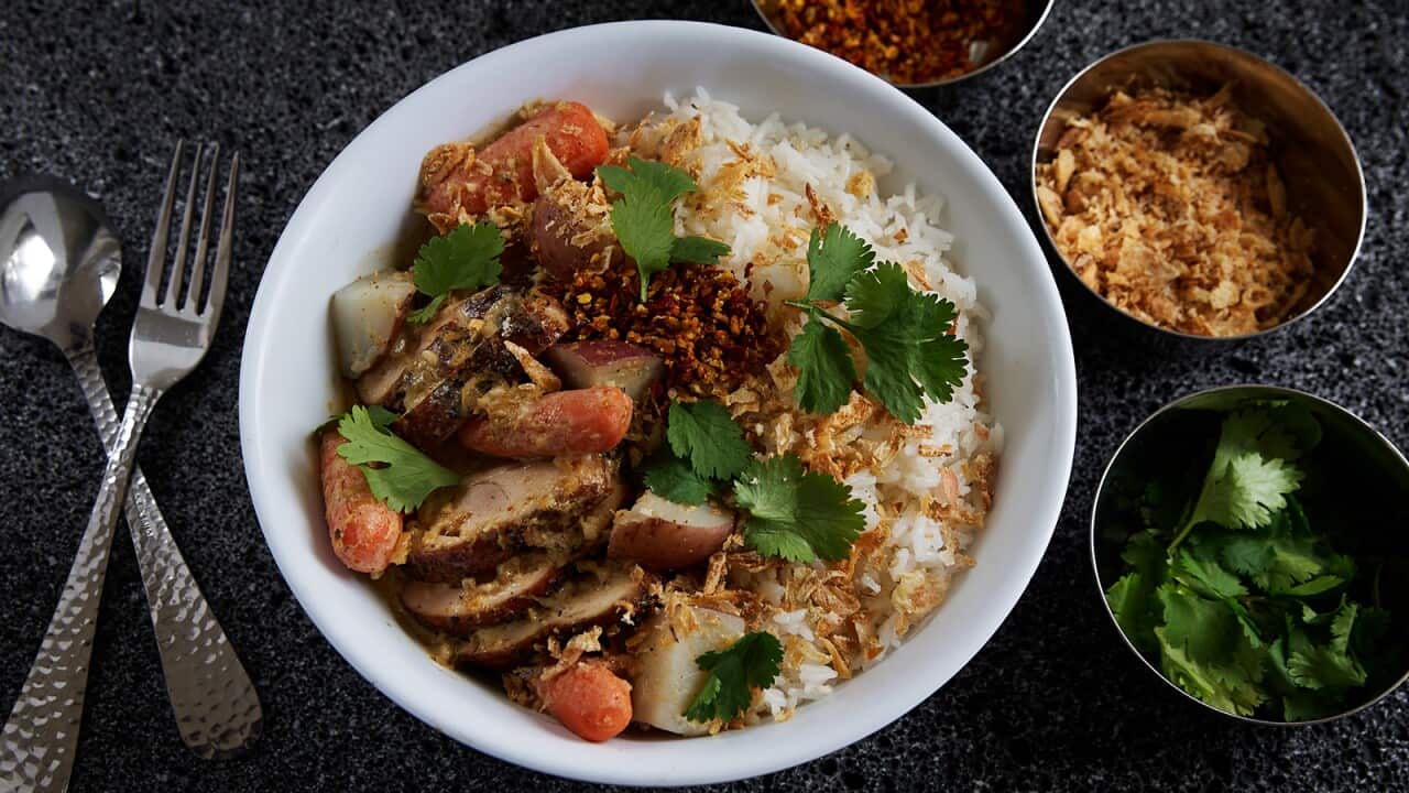 A white bowl sits on a dark surface, with a fork and spoon with textured handles alongside. The bowl holds rice, sliced chicken and vegetables, with scattered coriander leaves.