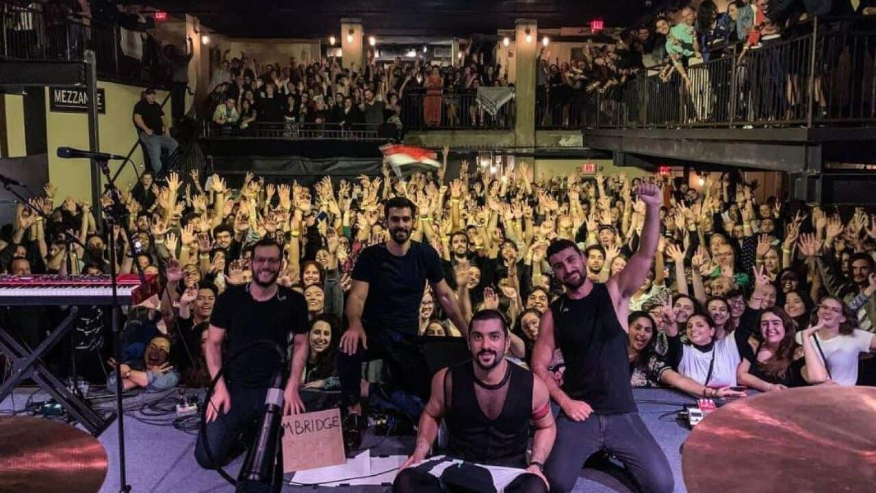 Four men dressed in black on a stage with hundreds of fans behind them.