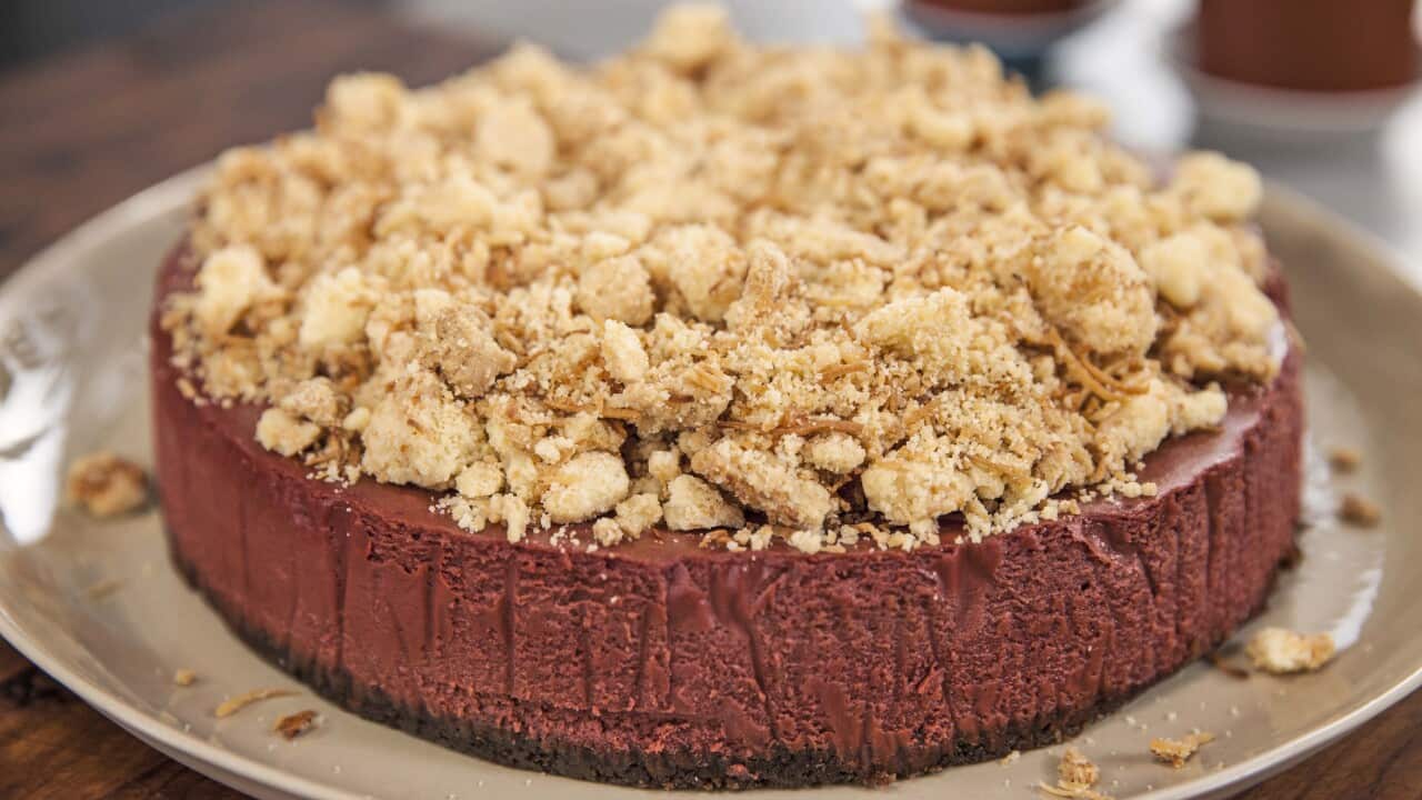 A reddish-brown cheesecake, topped with chunky golden crumbs, sits on a round plate.