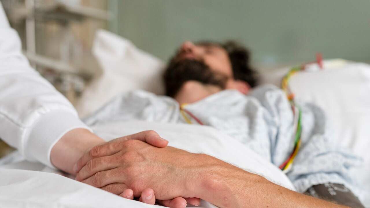 A man with a beard lies in a hospital bed, holding the hand of a healthcare worker
