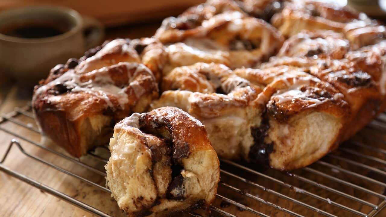 A rectangle-ish set of cooked sticky buns sits on a wire cooking rack, one is torn off the front and sites separately, showing the slighlty melted choc interior and sticky glaze.