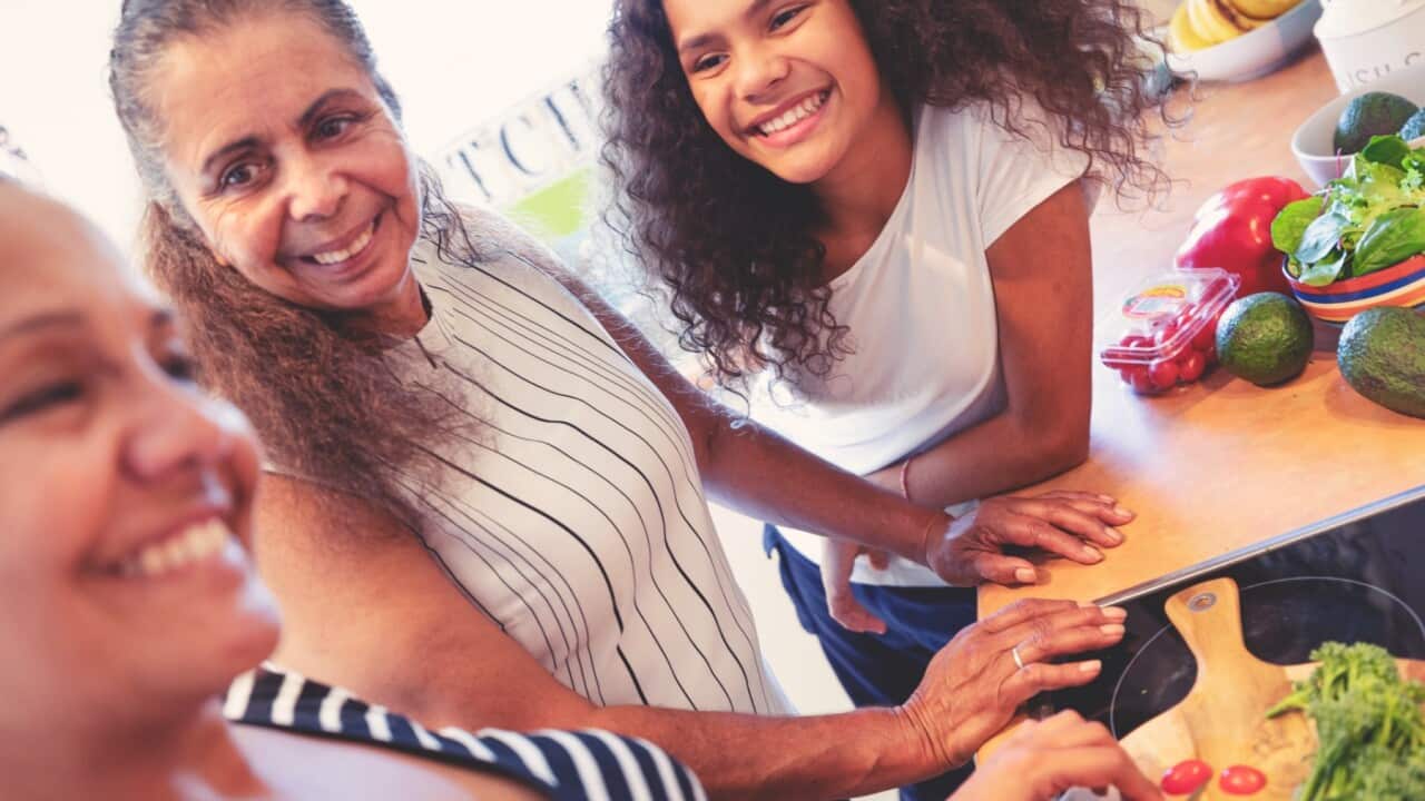Grandmother, Mother and child cooking in the kitchen. They are cooking a healthy meal and talking. Three generations. There are vegetables on a cutting board in the foreground including peppers, carrots, broccoli and tomatoes. They are having fun and are