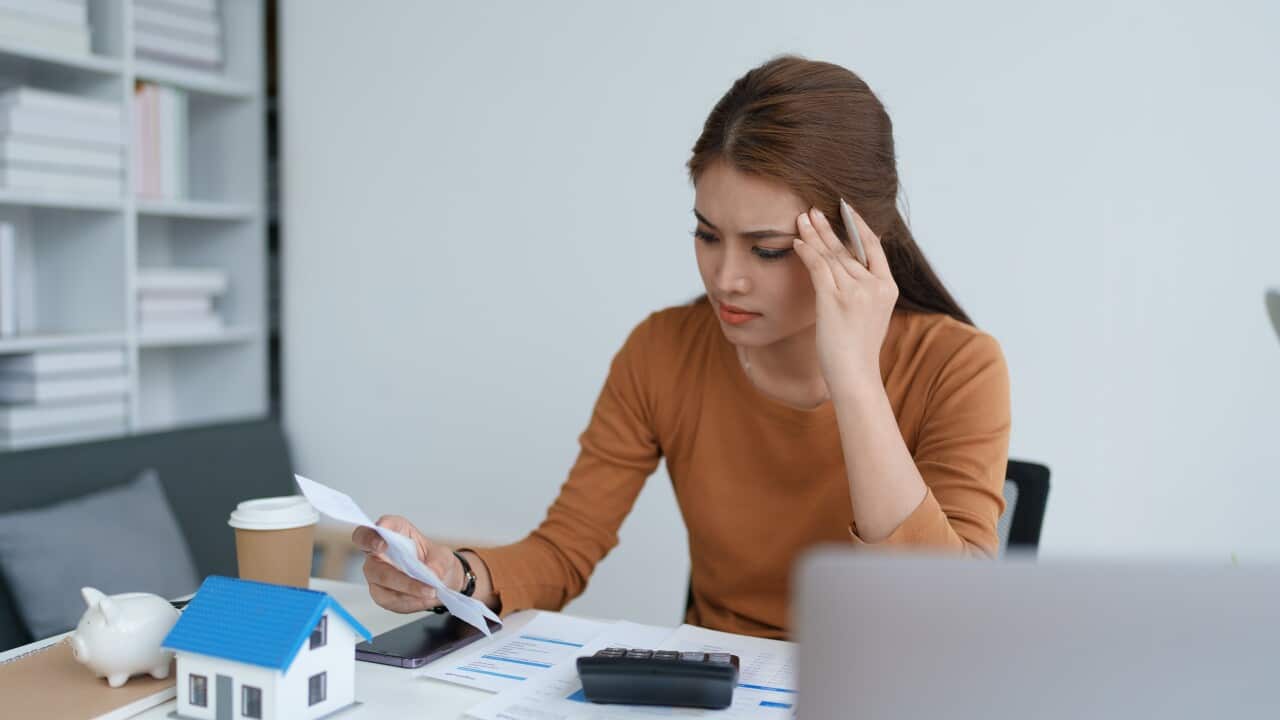 A stressed-looking woman sits at a table with sheets of paper, a laptop, a piggy bank and a model house in front of her.