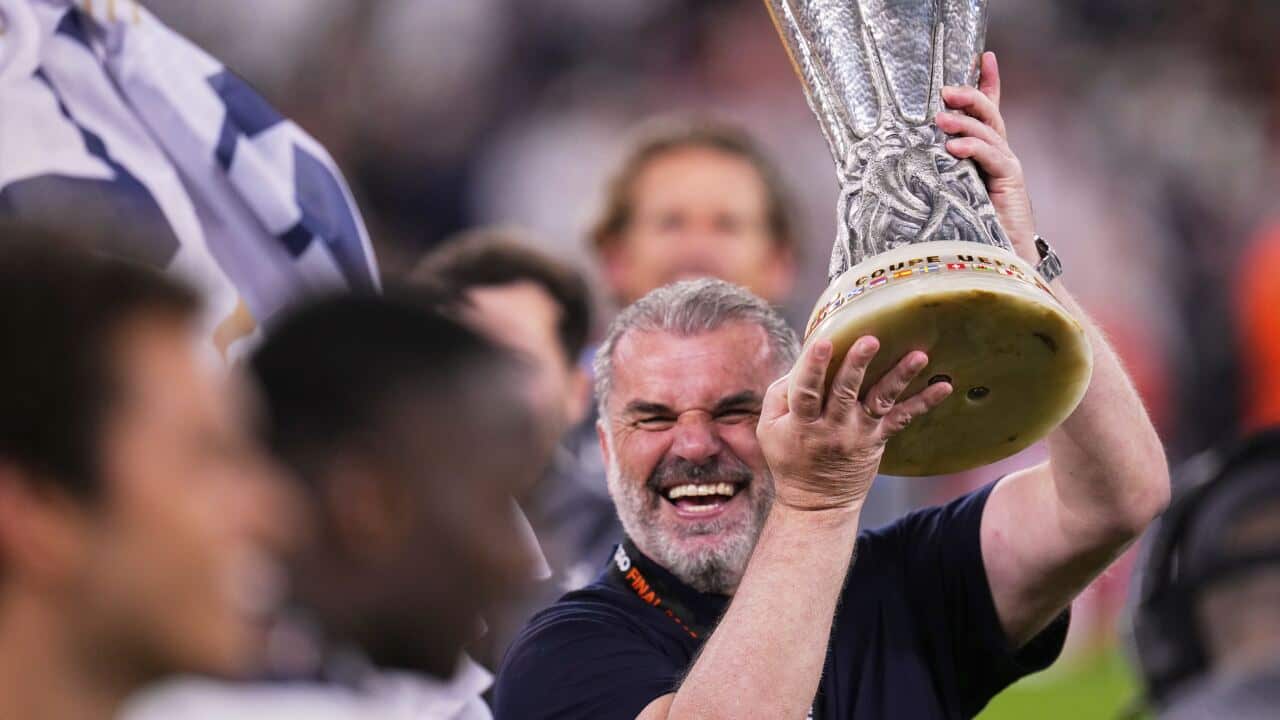 A man with grey hair and wearing a blue t-shirt is grinning as he holds up a trophy in his hands.