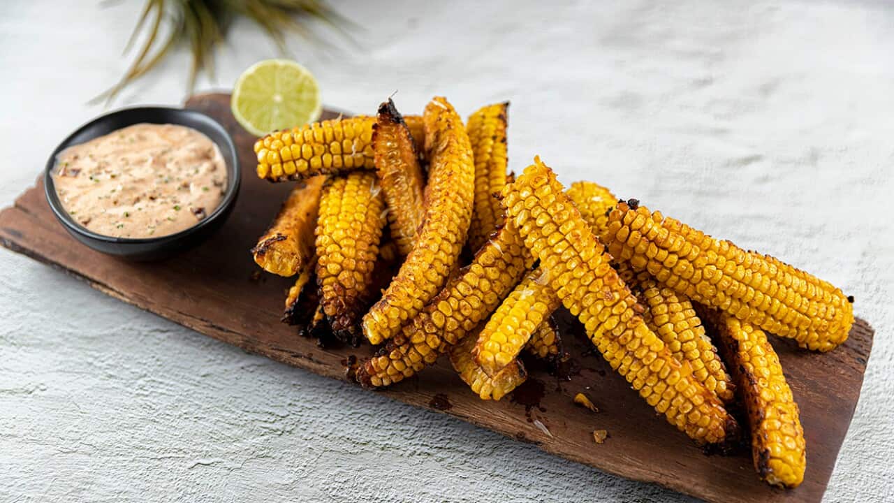 Long pieces of cooked corn sit on a long wooden board, along with a bowl of sauce and a piece of lime.