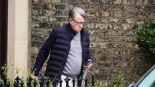 An older man wearing glasses and a dark jacket walks past a brick wall while carrying a folded umbrella and phone.