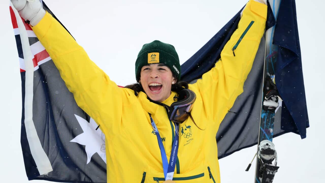 A woman wearing a yellow Australian jacket and a green beanie beams as she raises an Australian flag behind her. She's in a snowy area and clutching a ski in one hand, wearing a gold medal around her neck.