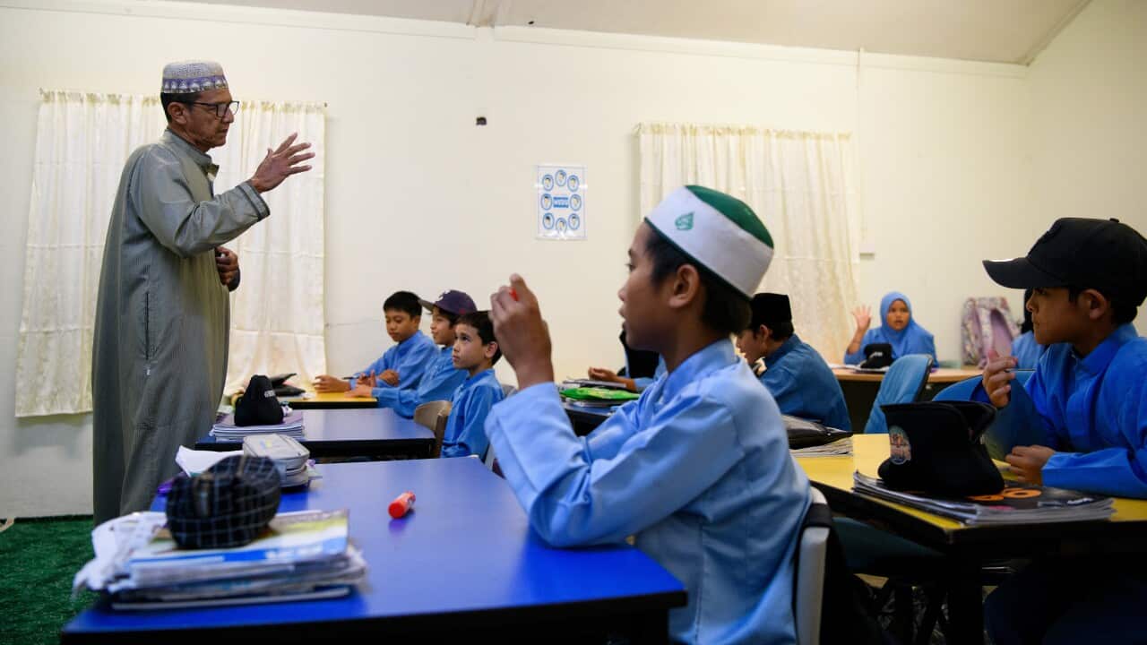 Children seated in a classroom listening to their teacher speak.