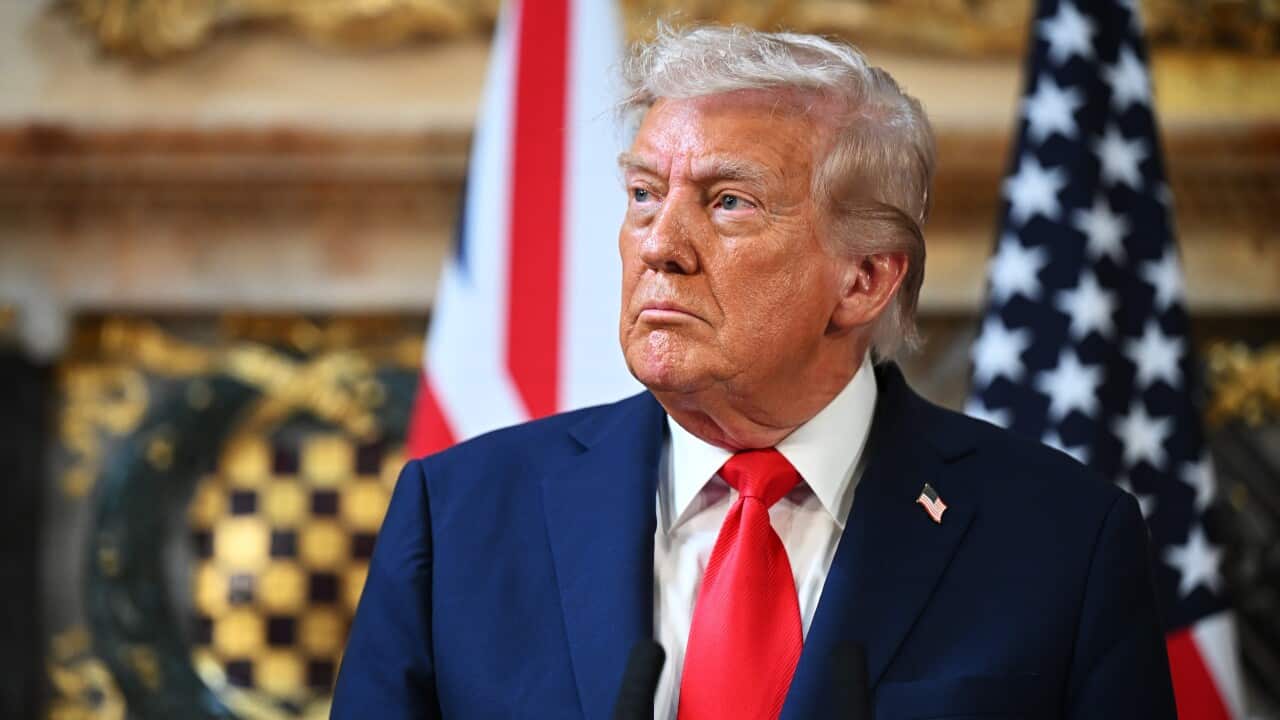 Donald Trump in a blue suit and red tie standing in front of two flags and a gold backdrop.