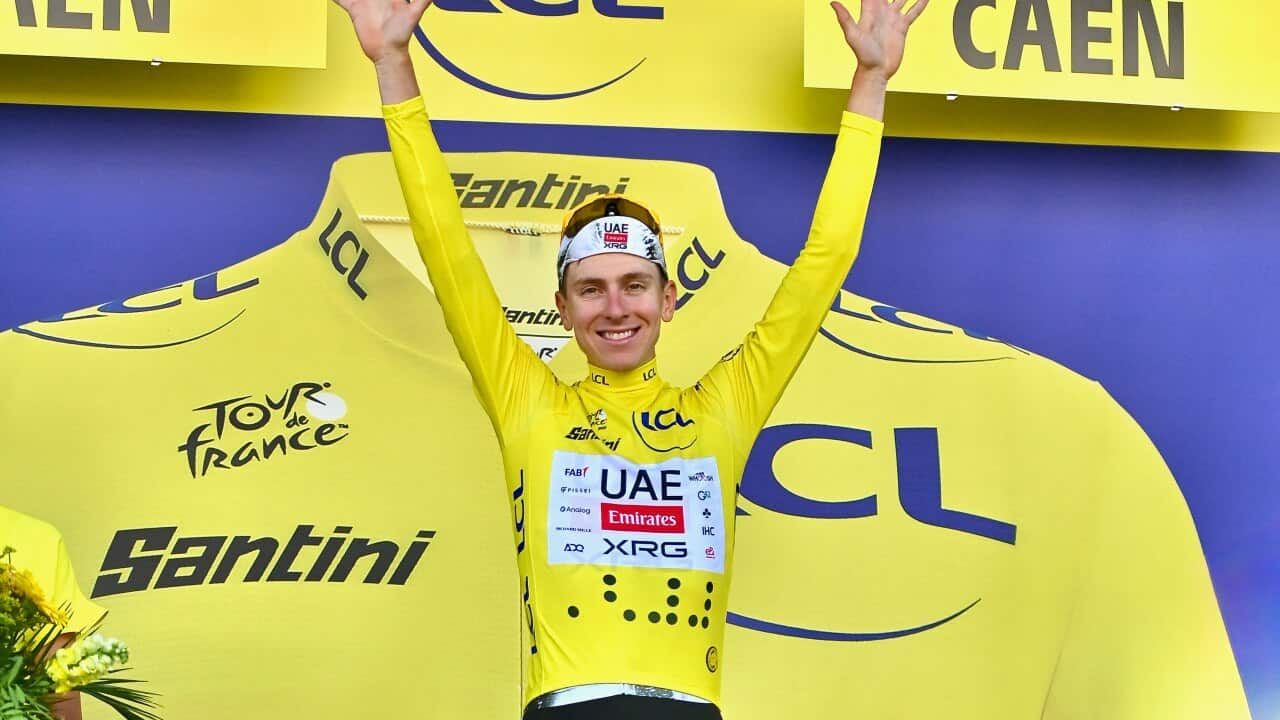 A man wearing a yellow jersey stands on a podium with his hands raised in victory. There's a screen displaying a yellow jersey behind him.