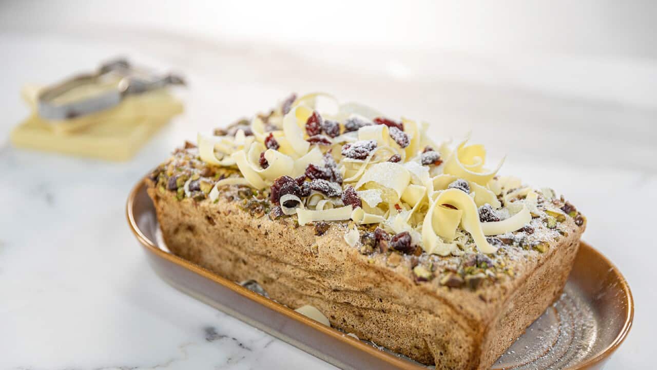 A loaf shaped cake sits on an oval serving dish. The cake is topped with pistachios, cranberries and white chocolate cruls.
