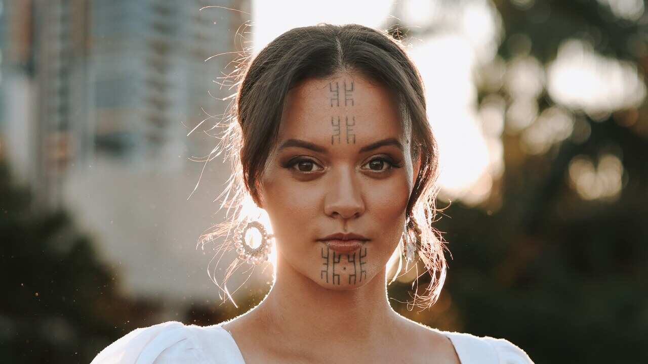 A woman with markings on her face looks at the camera with the sun behind her