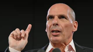 A close-up of Yanis Varoufakis speaking emphatically with his index finger raised against a dark background.