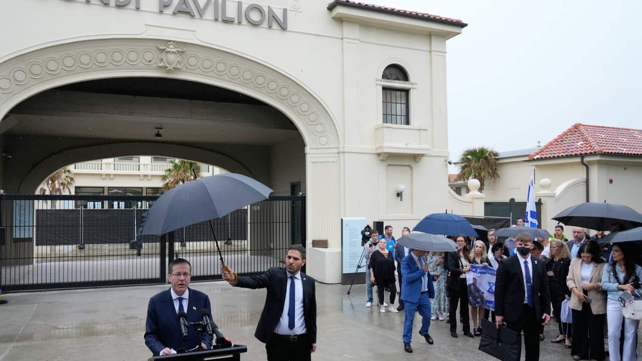 A man in a navy suit speaking before a lectern outside the Bondi Pavilion. To his left, people crowd under umbrellas as they watch on.