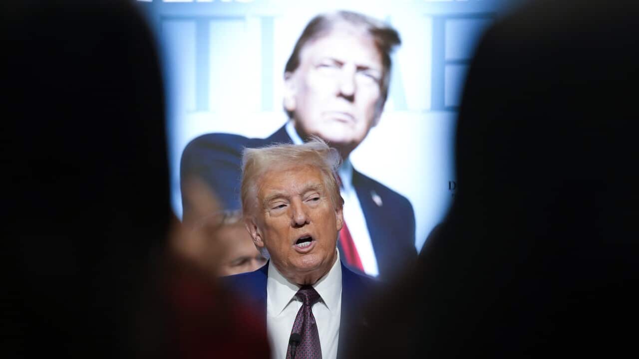 Man speaks in front of a poster of himself.