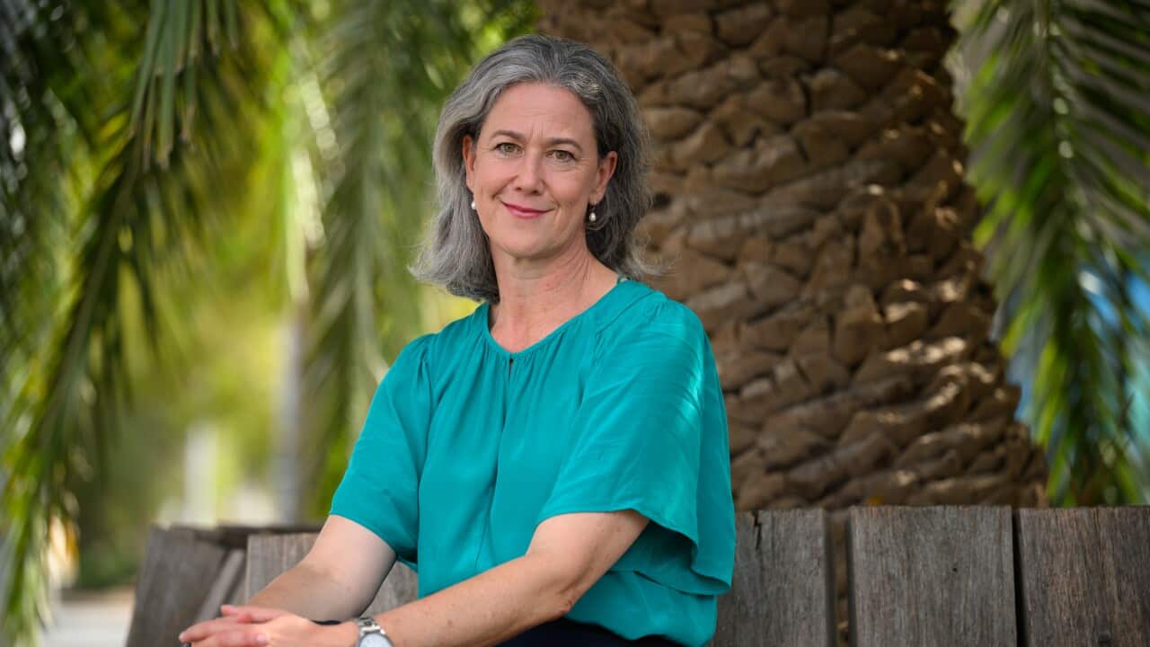 A woman in a turquoise shirt with silver hair is sitting on a wooden bench with her hands clasped on her knees.