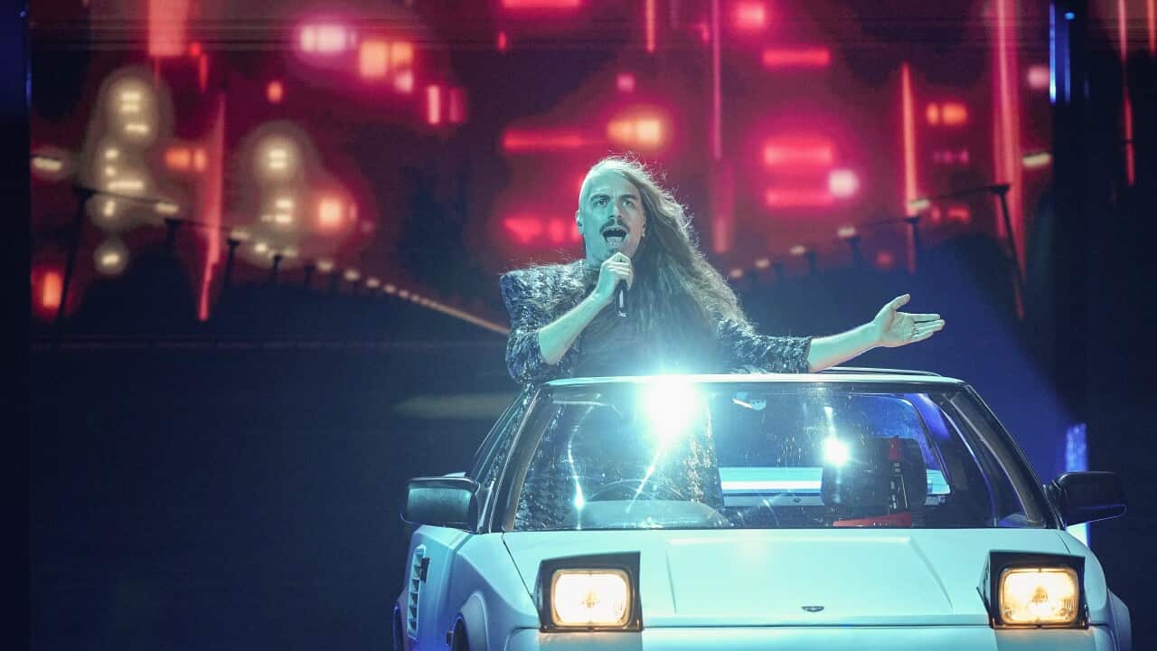A man with long hair singing in a car on a stage