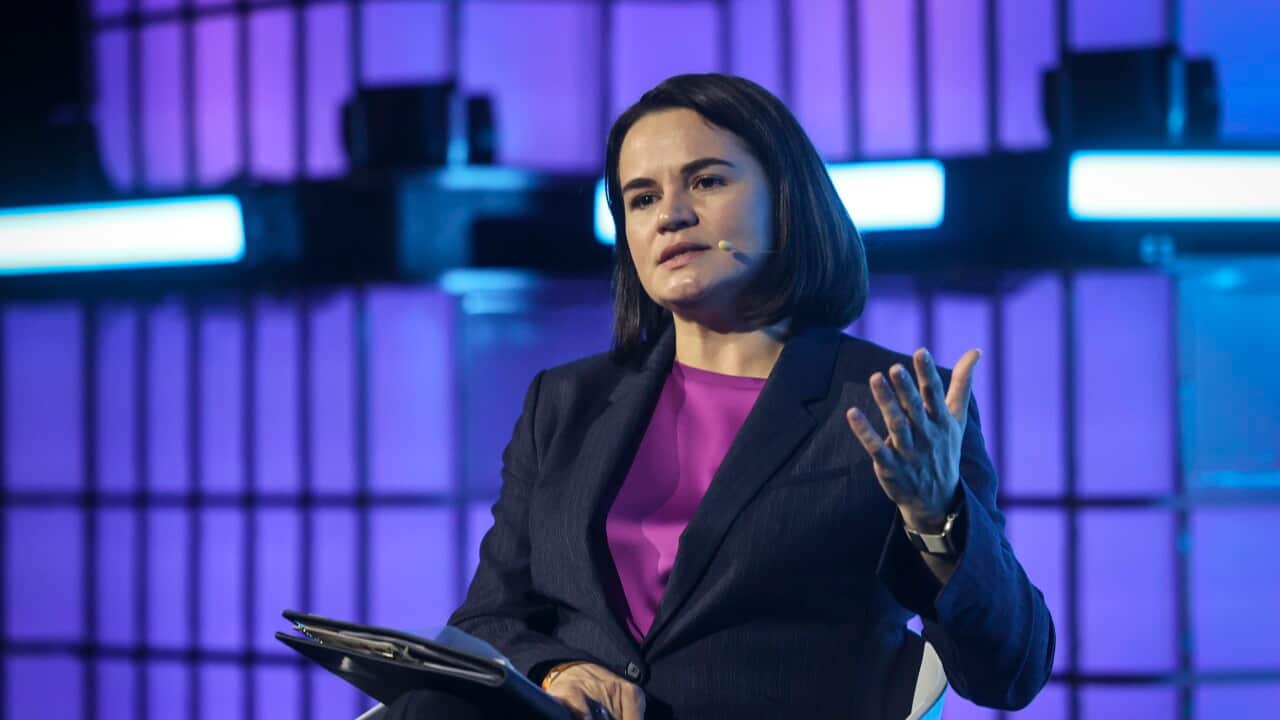 A woman wearing a dark suit and a bright purple top speaks while seated on a stage against a backlit grid.