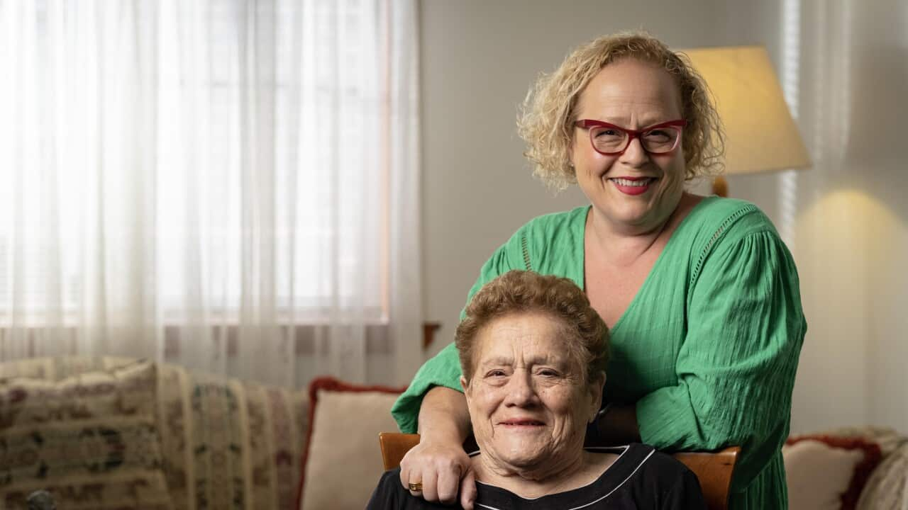 Image of a woman wearing black sitting down and a woman in a green dress and red glasses together in a cozy loungeroom.