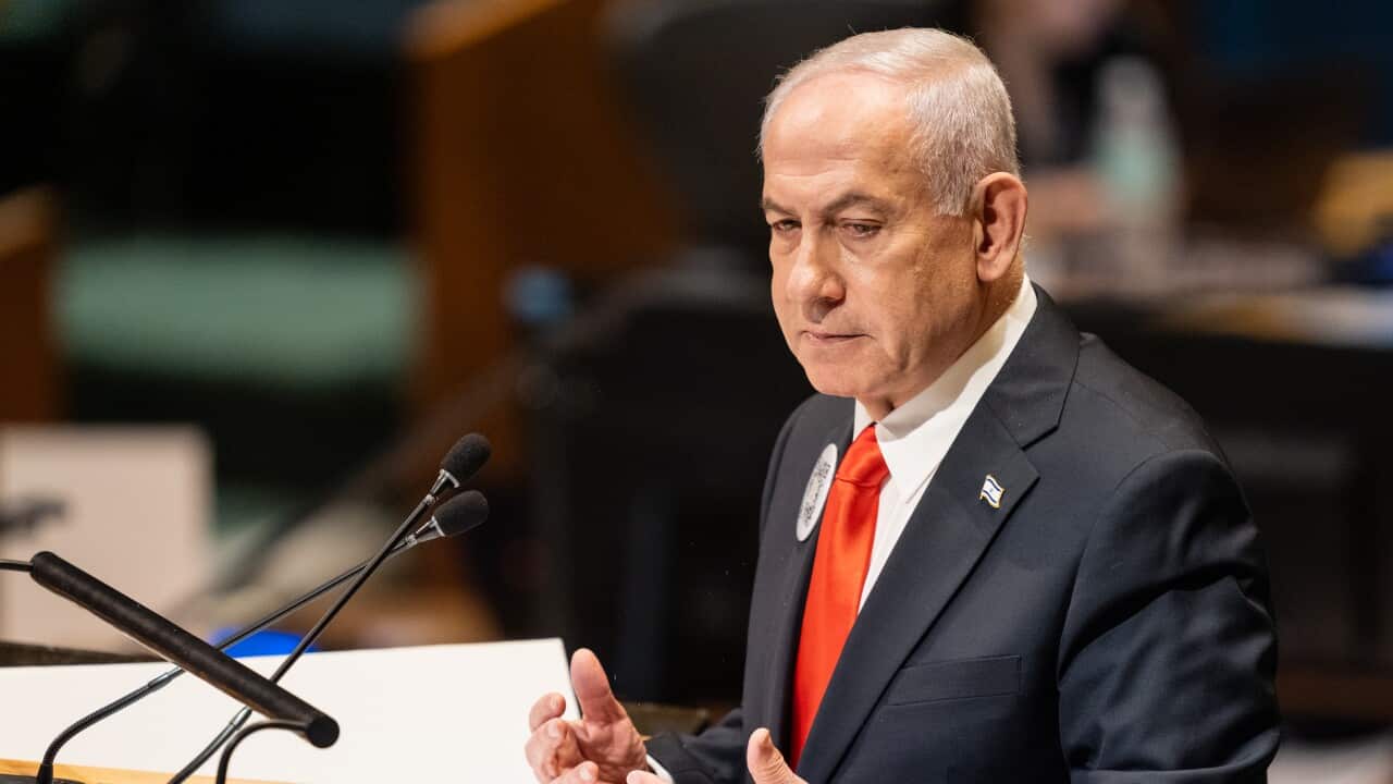 A close-up photograph shows Benjamin Netanyahu, dressed in a dark suit and a bright red tie, speaking at a podium with two microphones in front of him. He is mid-gesture, looking intently to his left, with a serious expression on his face in a dimly lit, official setting.