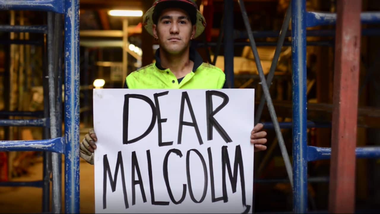a man in an orange vest and helmet on a work site holds a sign reading 'Dear Malcolm'