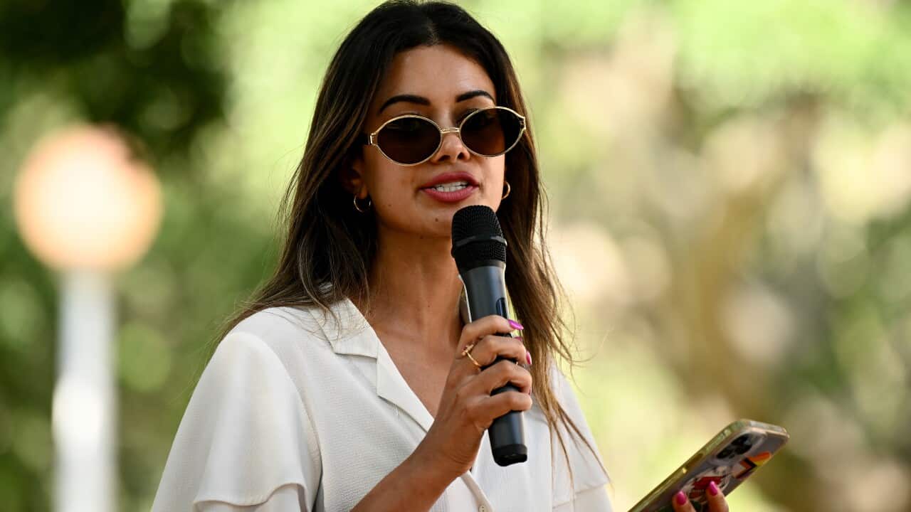 A woman in a white shirt wearing sunglasses and holding a phone and microphone in front of trees