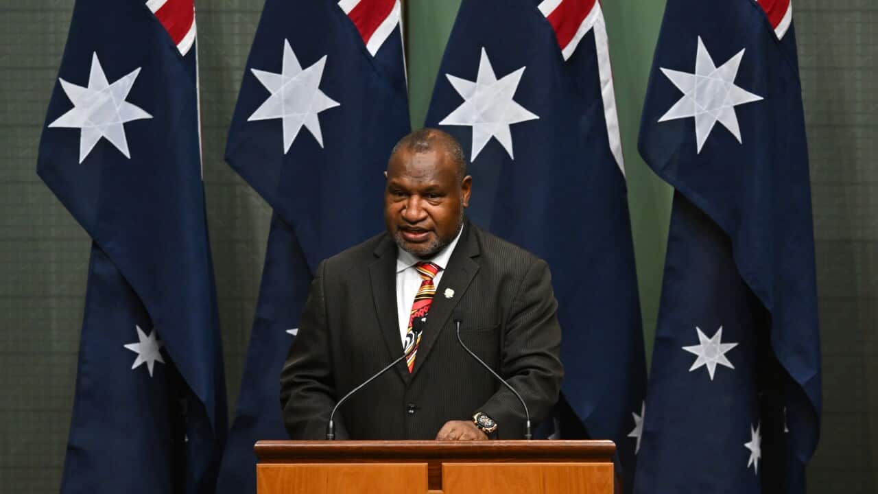 Man stands at wooden podium with two microphones attached, wearing a suit. There are four blue flags behind him, adorned with stars.