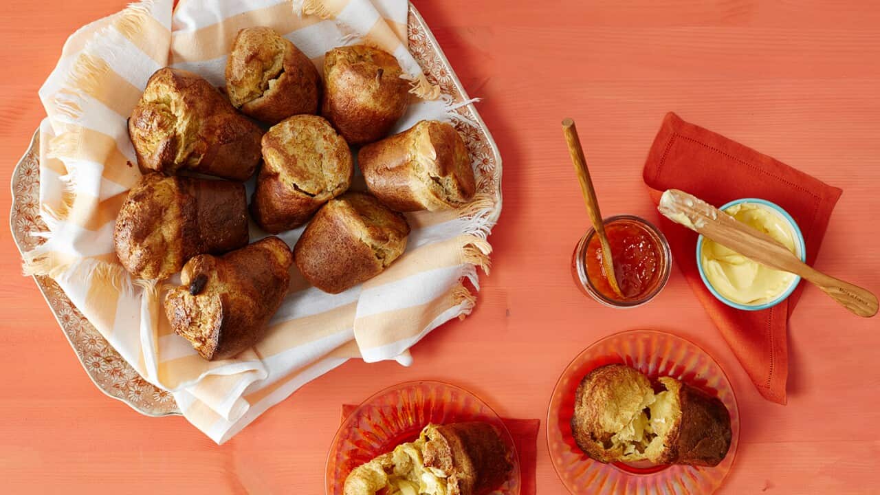 Seen from overhad, 8 golden-brown popovers, shaped like flower pots, sit on an apricot-and-white-striped napkin, on a floral plate. The plate,plus two torn-open  popovers on small plates, a jam pot and some butter, all sit on an orange surface.
