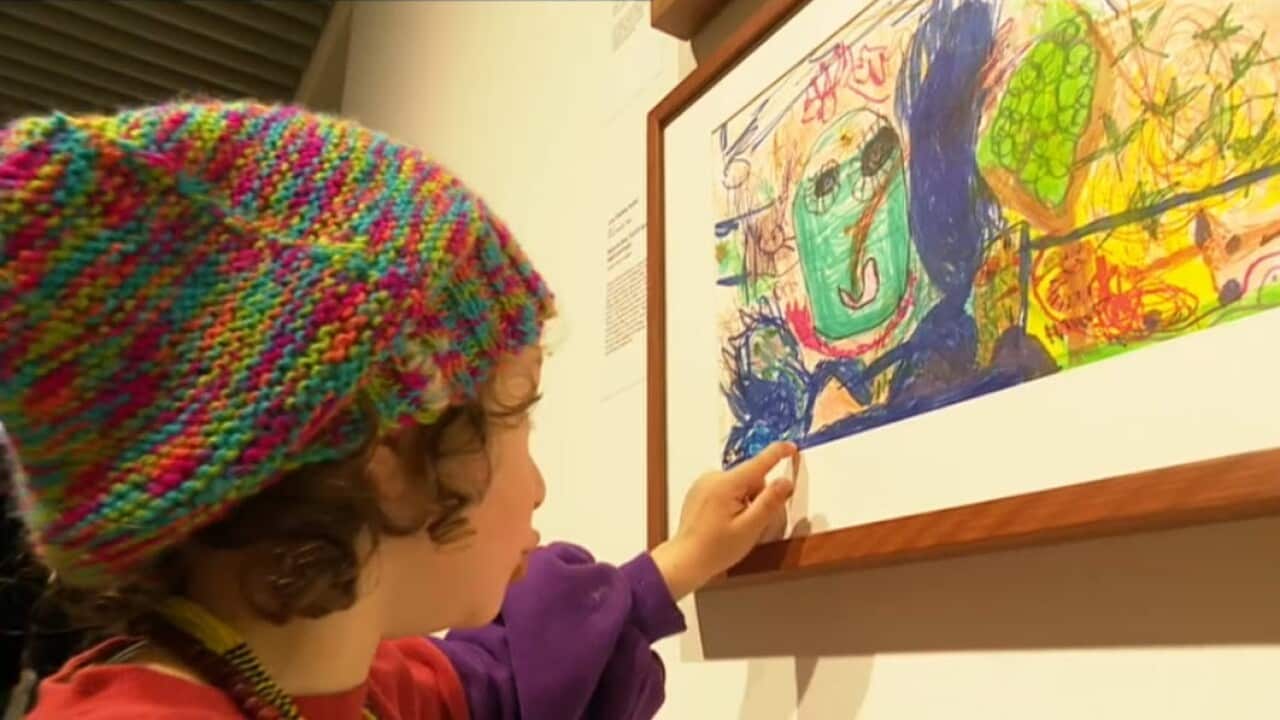 A young child in a rainbow beanie points to a colourful self-portrait they drew