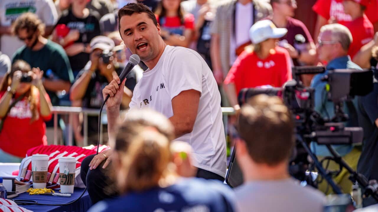 A man wearing a white shirt speaking into a microphone. Audience members surround him.