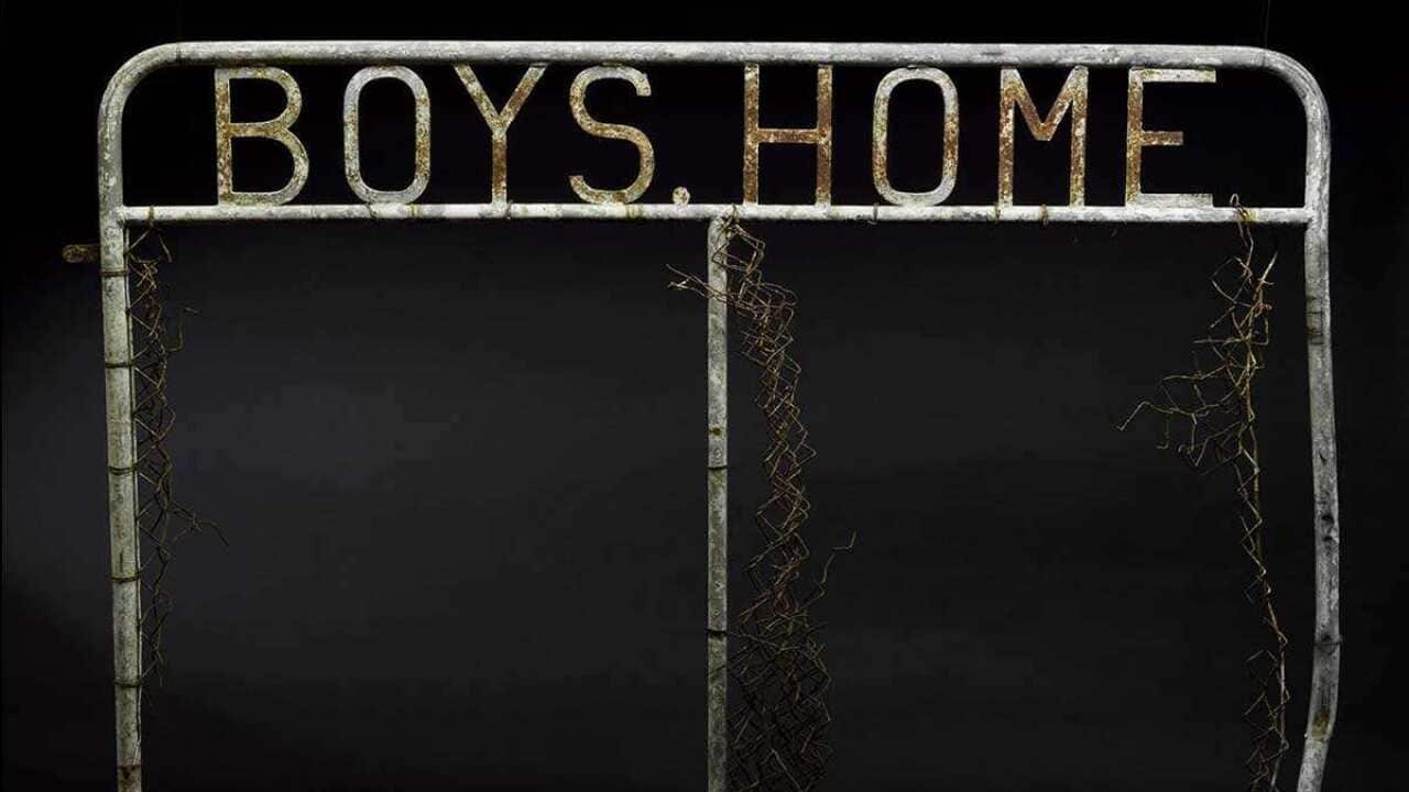 A rusted gate with the words "BOYS HOME" in cast-metal lettering. The wire mesh part of the gate has mostly fallen off.