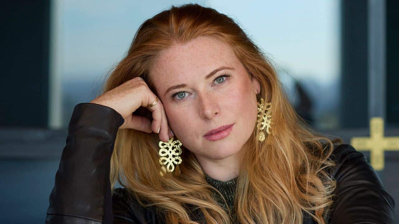 a closeup shot of a woman with long red hair wearing gold jewellery with her head resting on her hand
