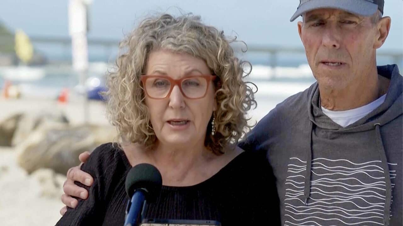 The father of the murdered Australian surfers wraps his arm around his wife as she speaks in front of a microphone.
