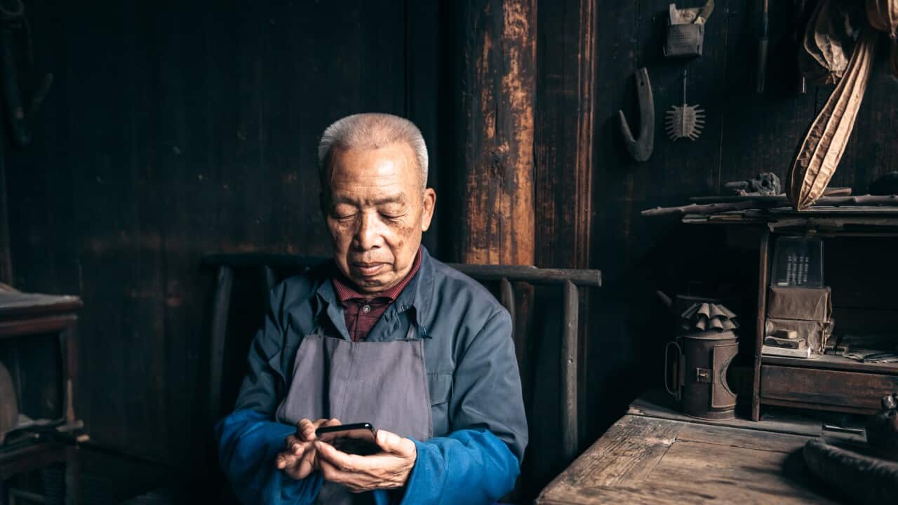 An elderly man in a workshop setting, wearing a blue shirt and gray apron, sits focused on his smartphone against a backdrop of traditional wooden architecture and tools.