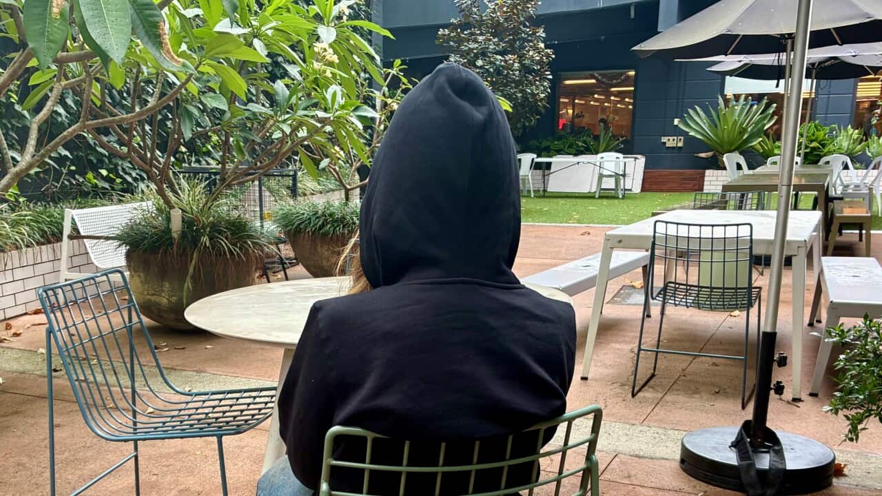 A person wearing a black hooded jacket sits in a courtyard.