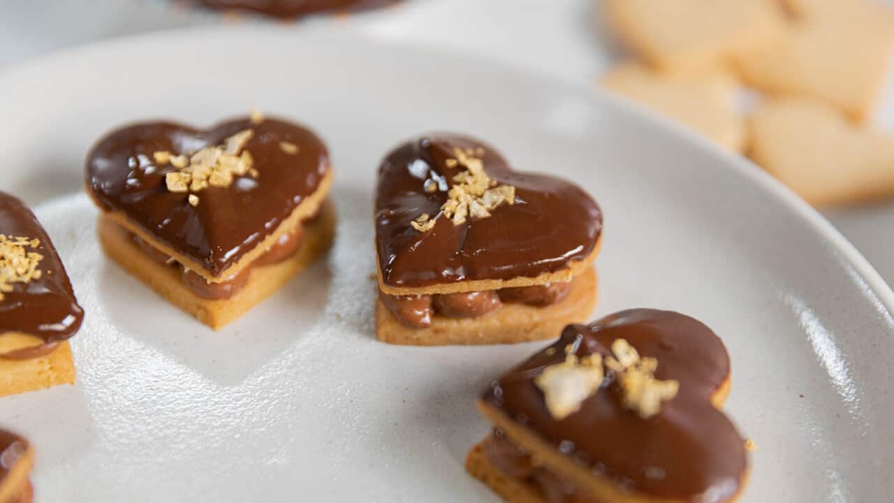 A ring of filled heart-shaped biscuits sit on a white plate. Each two-biscuit stack has a piped milk chocolate covered filling and a glossy dark chocolate icing on top.