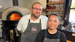 A man in a white apron and a woman in a black t-shirt stand in front of a wood-fired pizza oven. 