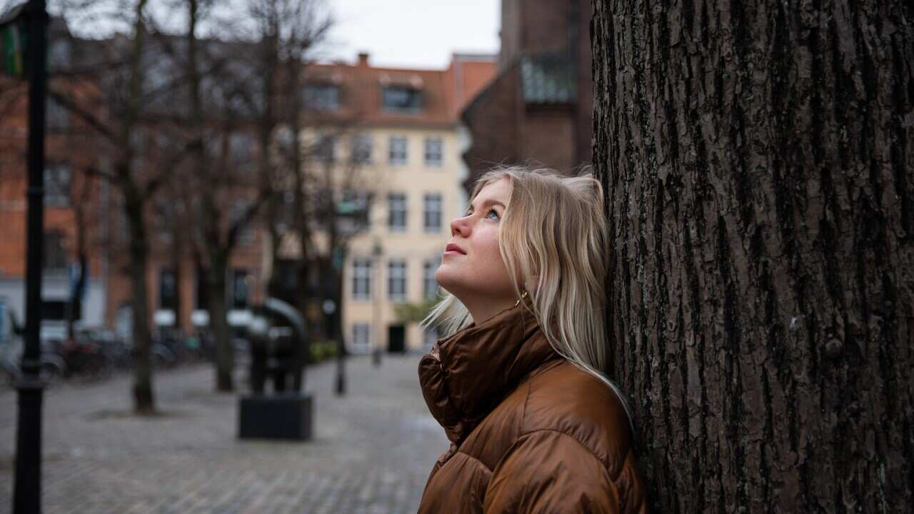 A woman with blonde hair leans up against a tree.