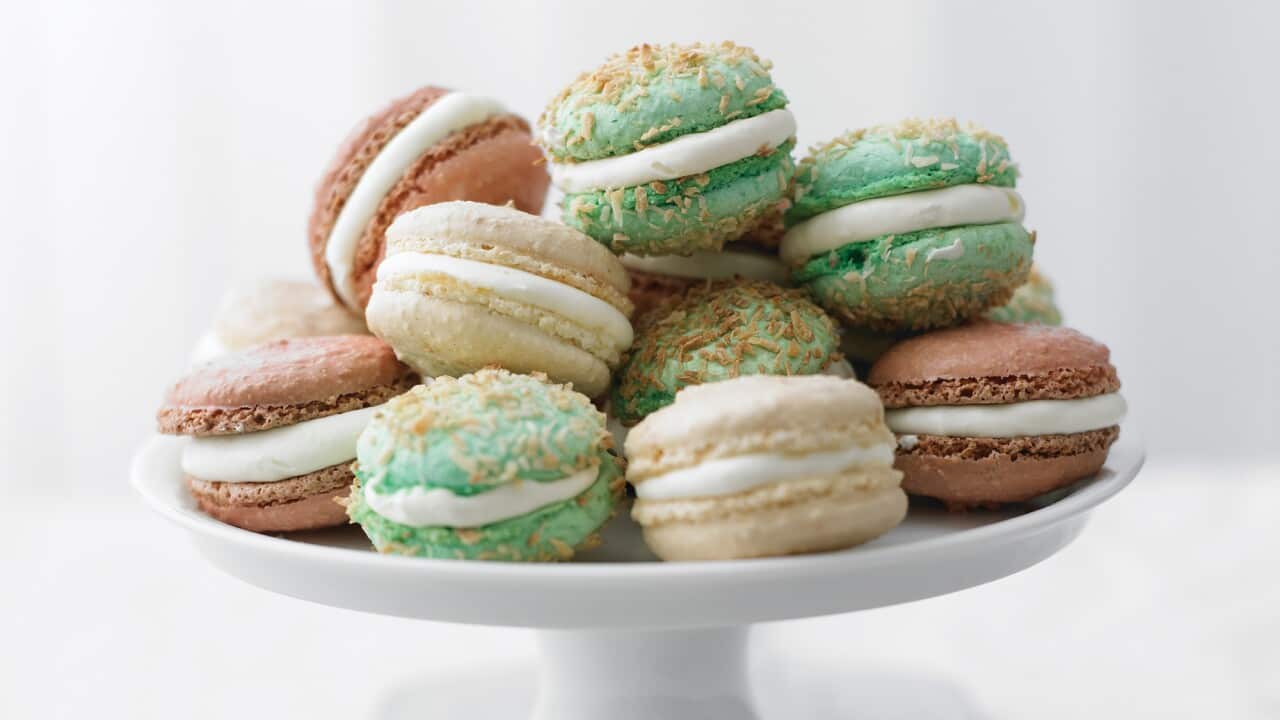Cream, pink and green-tinted macarons, sandwiched with cream, sit on a white cake stand.