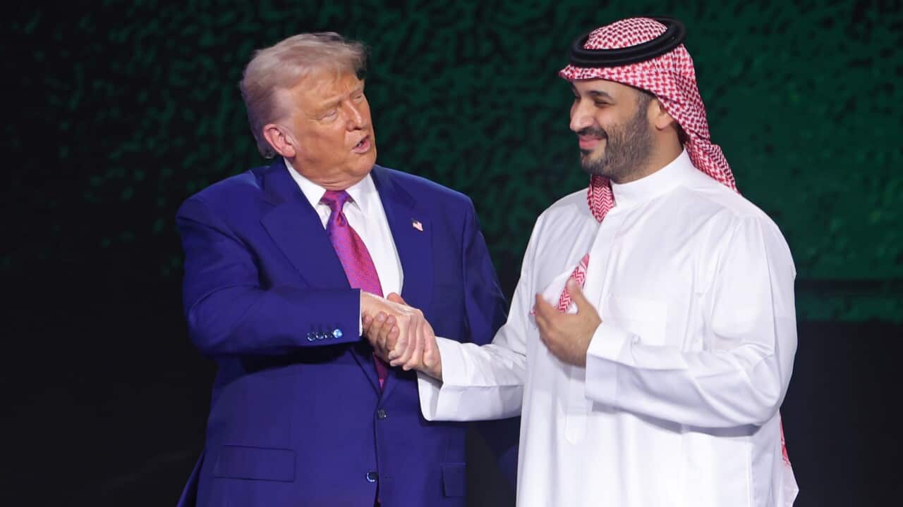 A man in a blue suit on the left shakes hands with a man in a white thobe and keffiyeh on his head.