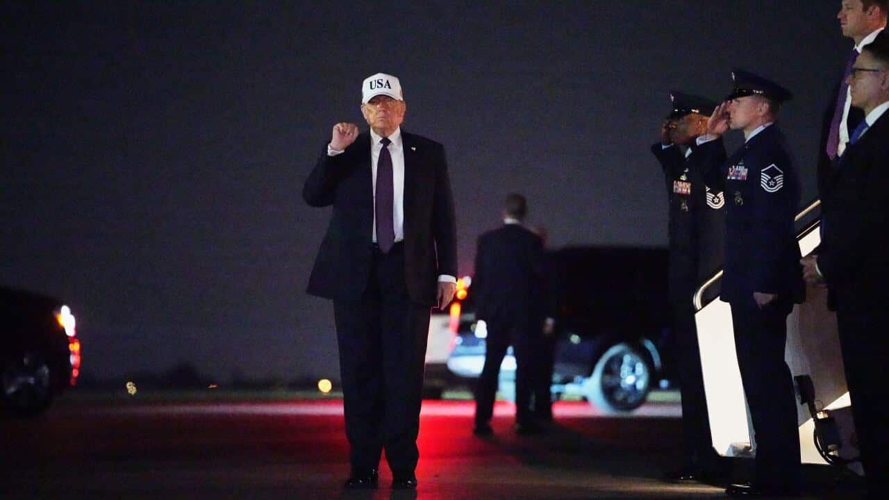 Donald Trump, wearing a white “USA“ hat and a dark suit, raises a fist while walking on a tarmac at night as military personnel salute in the background.