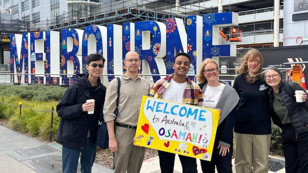 A man holding a hand-drawn 'welcome' sign, surrounded by a group of people standing in front of a large sign that says 'Melbourne.'
