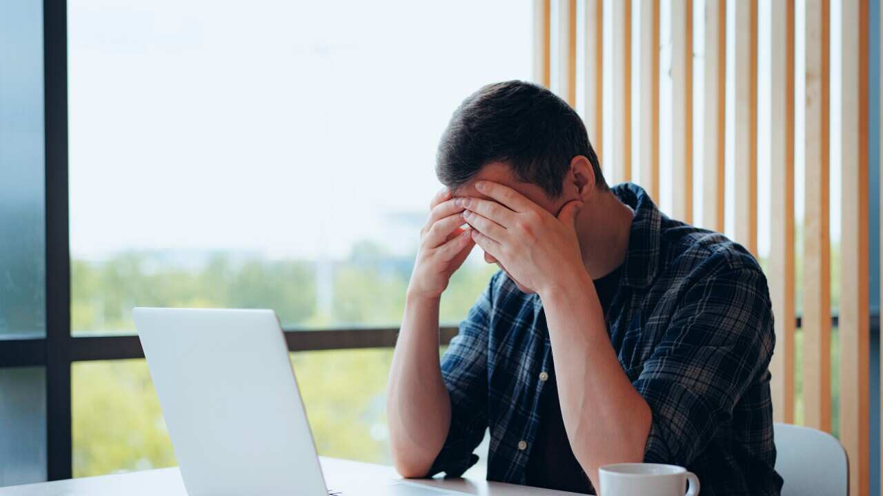 A man wearing a blue tartan shirt, sitting in front of a laptop with his head in his hands
