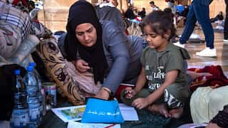 A woman wearing a headscarf reaches into a pencil case with Arabic writing on it. A young girl is sitting next to her and several sheets of paper with drawings on them sit in front of them. Several bottles of water and a small portable gas stove are next to them.