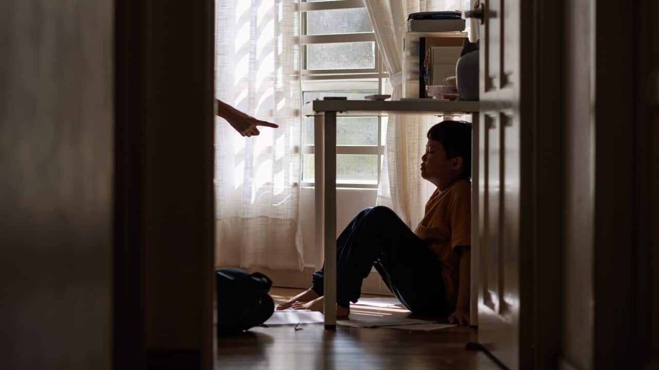 A little Asian boy with a sad and stress expression hides in the shadows under a table at home, while being scolded by his mother