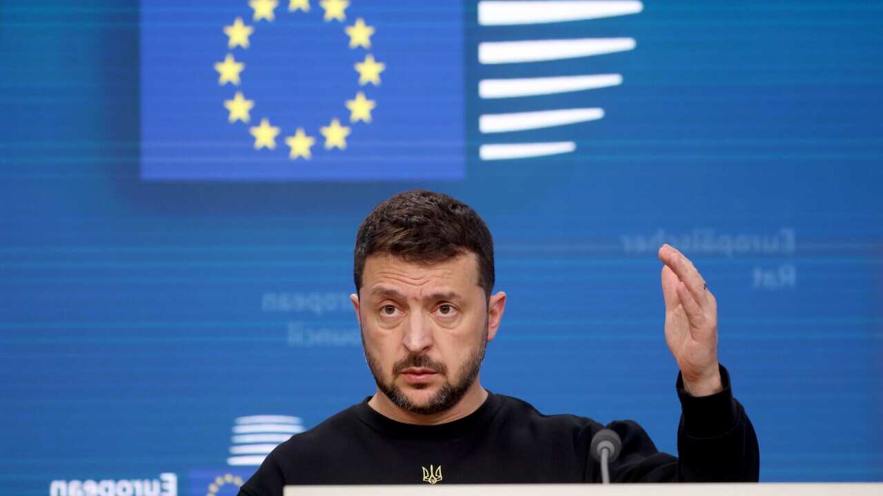 Volodmyr Zelenskyy standing behind lectern. There is a blue background behind him and he is holding up his left hand.